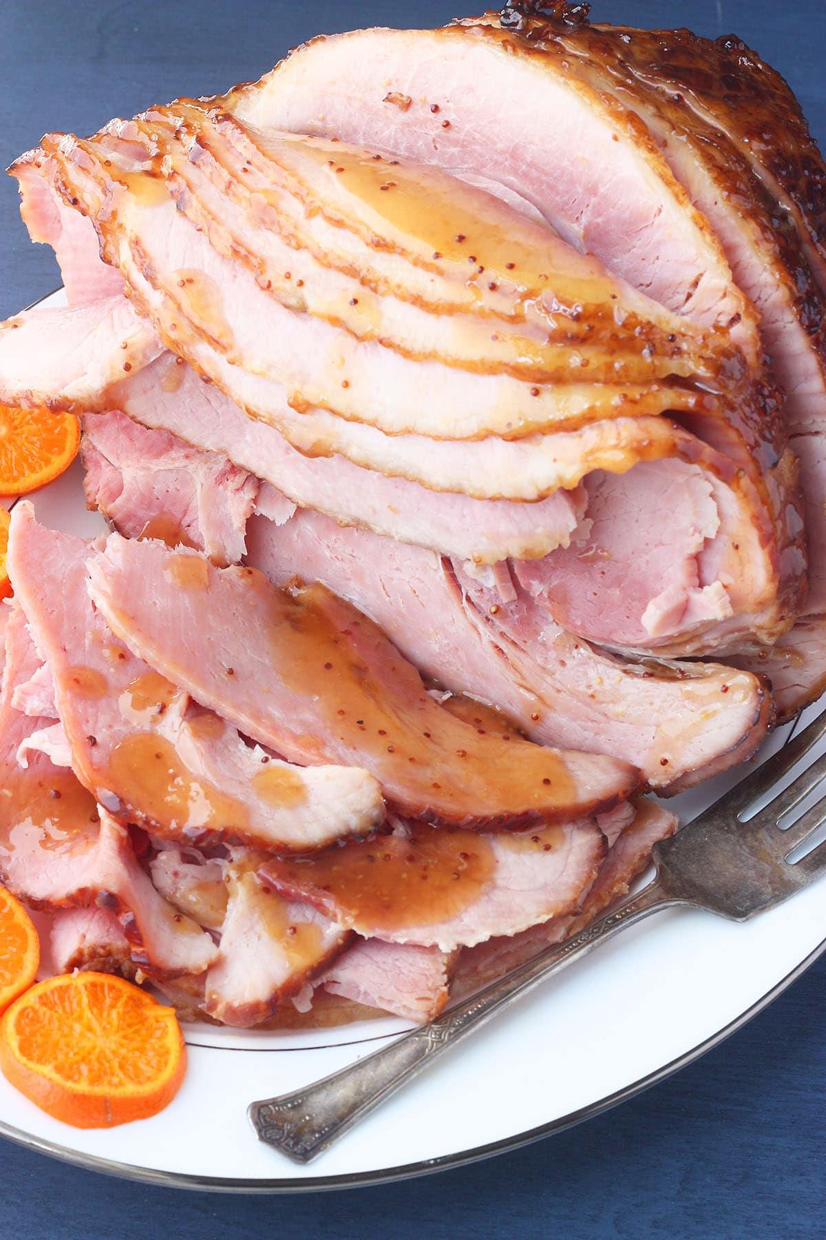 side shot of sliced bourbon glazed ham on a white platter with sliced oranges on the side