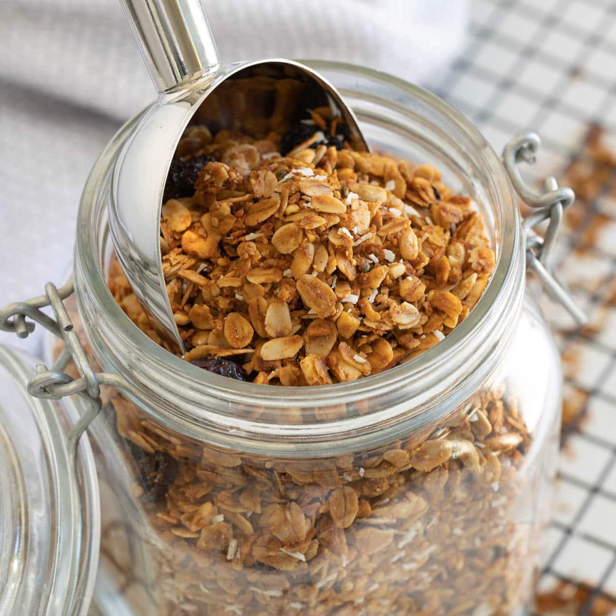A close up of an open jar of homemade granola with a silver scoop.